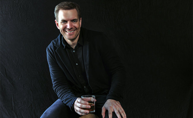Portrait photo of Brian Dickett, smiling in a dark jacket while seated against a black backdrop and holding a glass of whiskey.
