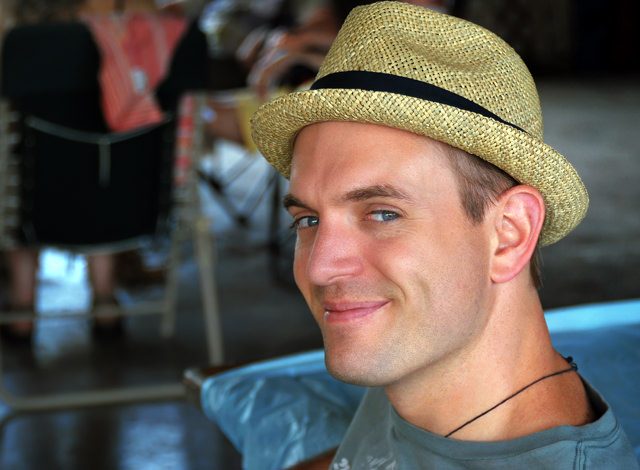 Portrait of Brian Dickett, smiling in a straw hat and patterned shirt, standing outdoors in front of a bright, tropical-looking background.