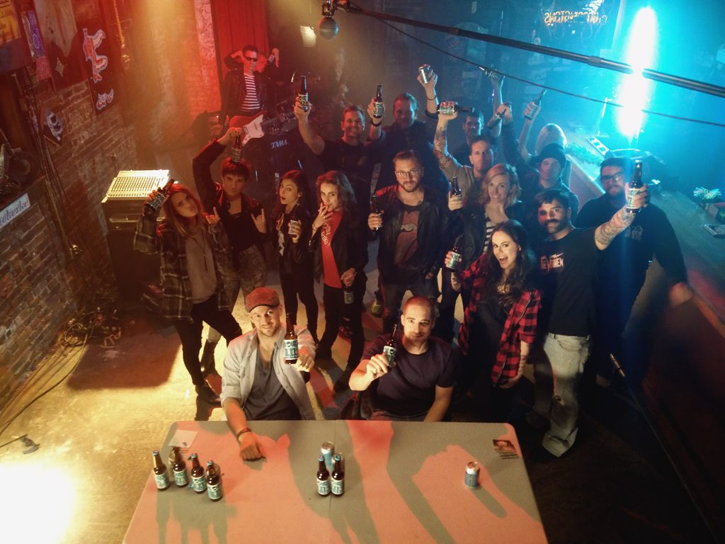 Behind-the-scenes group photo from from the BrewDog Punk IPA shoot, with cast and crew raising beer bottles of beer on a colorful bar set with stage lighting, instruments, and production equipment.