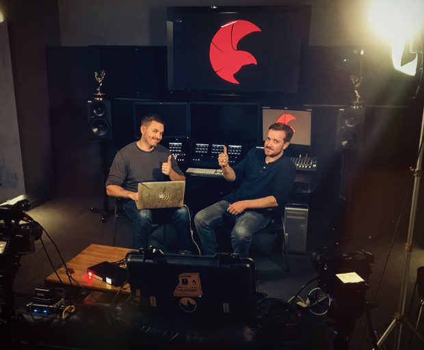 Behind-the-scenes view of Redtail Media Managing Partners, Brian Dickett and Chris Burke seated in a video production studio, giving thumbs up beside cameras, lighting equipment, monitors, speakers, and a large screen with the Redtail Media logo.
